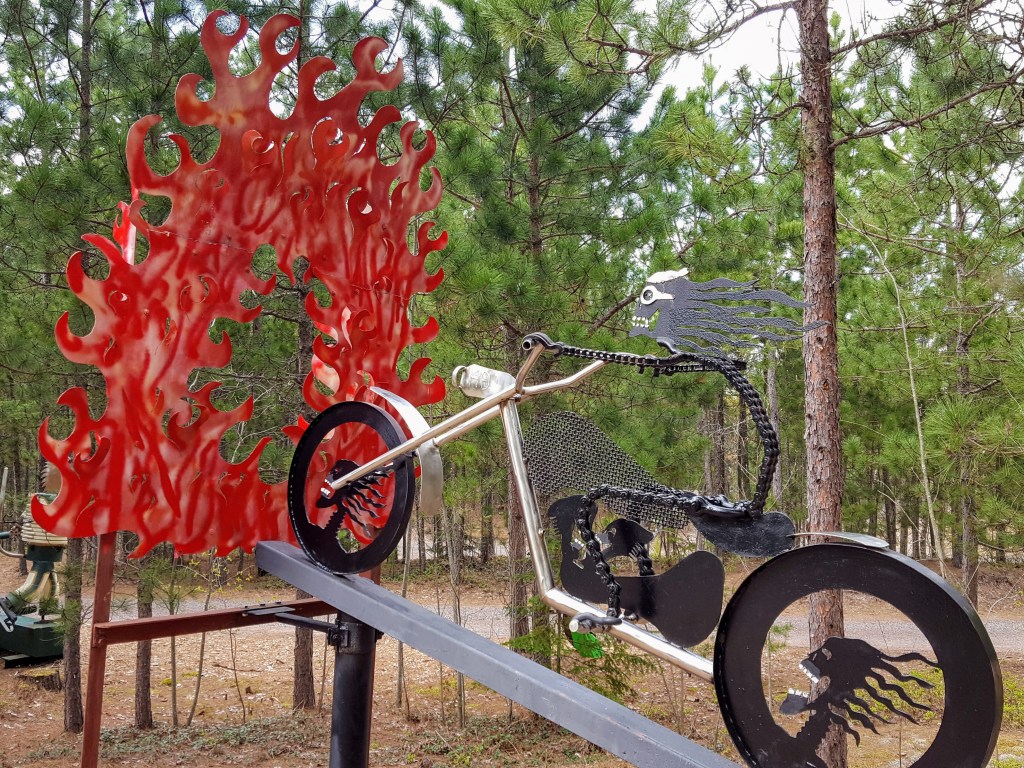 Picture a metal sculpture of a skeleton motorcyclist riding through a ring of fire in the Lakenenland Sculpture park.